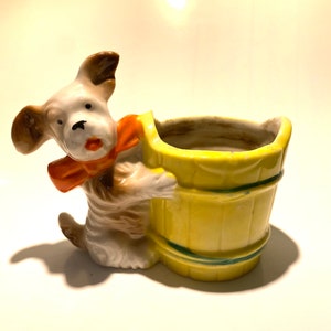 May include: A ceramic planter shaped like a dog holding a yellow bucket. The dog has brown ears, a white body, and an orange bow tie.