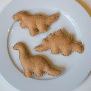 Pu&ograve; includere: Tre giocattoli di dinosauro in feltro marrone su un piatto bianco. I dinosauri sono uno stegosauro, un brachiosauro e un tirannosauro rex.