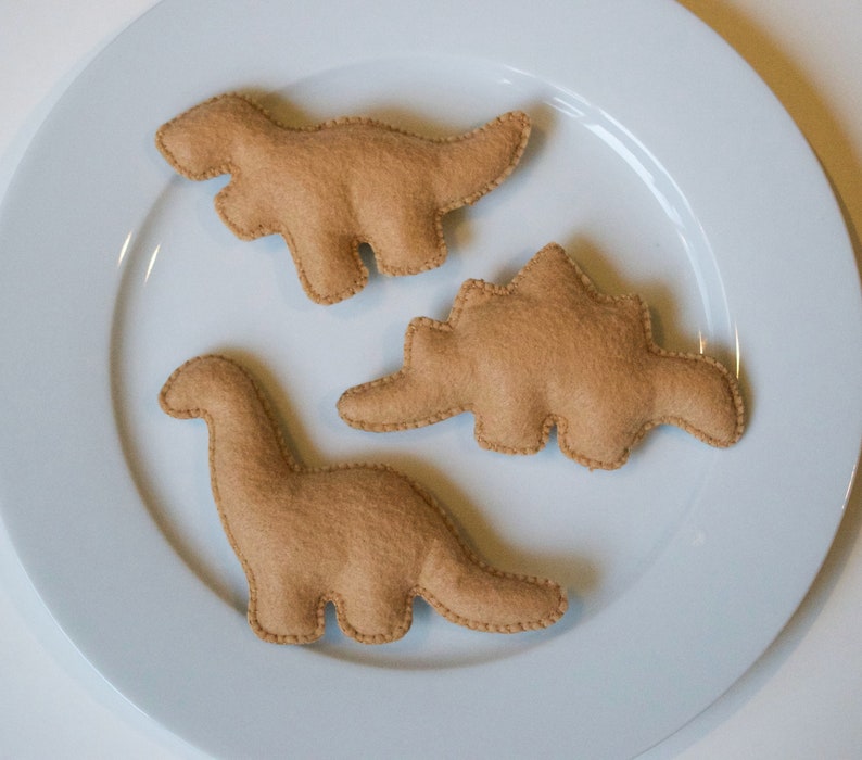 Pu&ograve; includere: Tre giocattoli di dinosauro in feltro marrone su un piatto bianco. I dinosauri sono uno stegosauro, un brachiosauro e un tirannosauro rex.
