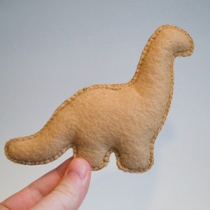 Pu&ograve; includere: Un giocattolo di dinosauro in feltro marrone con dettagli cuciti. Il dinosauro &egrave; in piedi sulle zampe posteriori con la coda arrotolata dietro di lui.