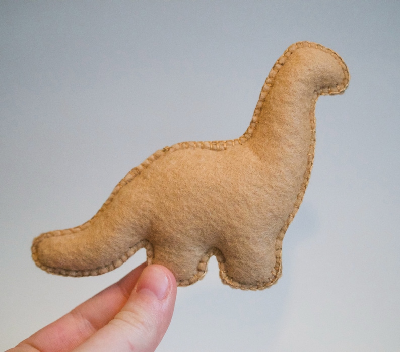 Pu&ograve; includere: Un giocattolo di dinosauro in feltro marrone con dettagli cuciti. Il dinosauro &egrave; in piedi sulle zampe posteriori con la coda arrotolata dietro di lui.