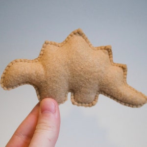 Pu&ograve; includere: Un giocattolo di dinosauro in feltro marrone con un contorno cucito. Il dinosauro ha la forma di uno stegosauro con una coda lunga e una fila di piastre sulla schiena.
