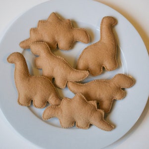 Pu&ograve; includere: Sei forme di dinosauro in feltro marrone su un piatto bianco. I dinosauri sono di diverse dimensioni e forme, e sono tutti rivolti in direzioni diverse.