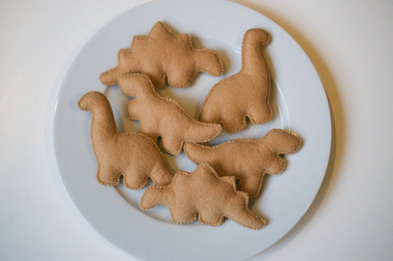 Pu&ograve; includere: Sei forme di dinosauro in feltro marrone su un piatto bianco. I dinosauri sono di diverse dimensioni e forme, e sono tutti rivolti in direzioni diverse.