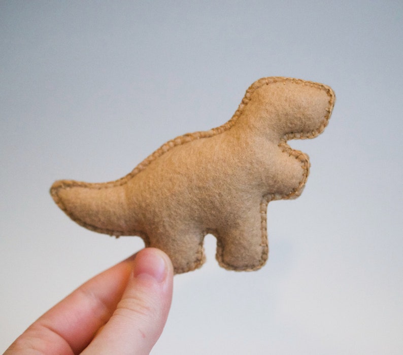 Pu&ograve; includere: Un giocattolo di dinosauro in feltro marrone. Il dinosauro &egrave; in piedi sulle zampe posteriori con la coda arrotolata dietro di lui.