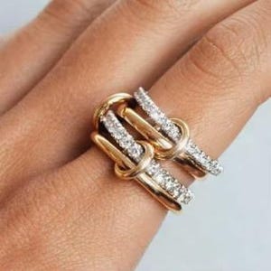 Puede incluir: Un anillo dorado con diseño de nudo, con dos bandas entrelazadas. Cada banda está adornada con una fila de pequeñas piedras brillantes. El anillo se muestra en un dedo, destacando sus intrincados detalles y su estilo elegante.