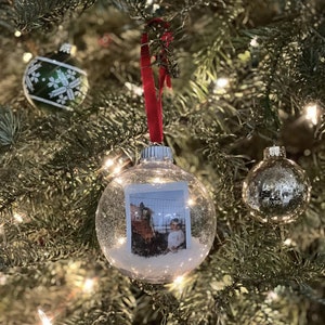 Peut inclure: Une boule de Noël en verre transparente avec un effet de neige scintillante blanche. À l'intérieur de la boule se trouve une photo d'un enfant et d'un cheval. La boule est suspendue à un sapin de Noël avec un ruban rouge.