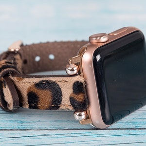 May include: A rose gold Apple Watch with a leopard print leather band. The band has a brown leather buckle and silver metal accents.