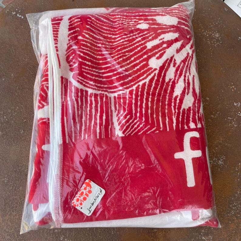 May include: A red and white beach towel, folded and packaged in clear plastic. The towel features a bold, abstract design in white against a red background. The letter "f" is visible in white. A small tag is attached.