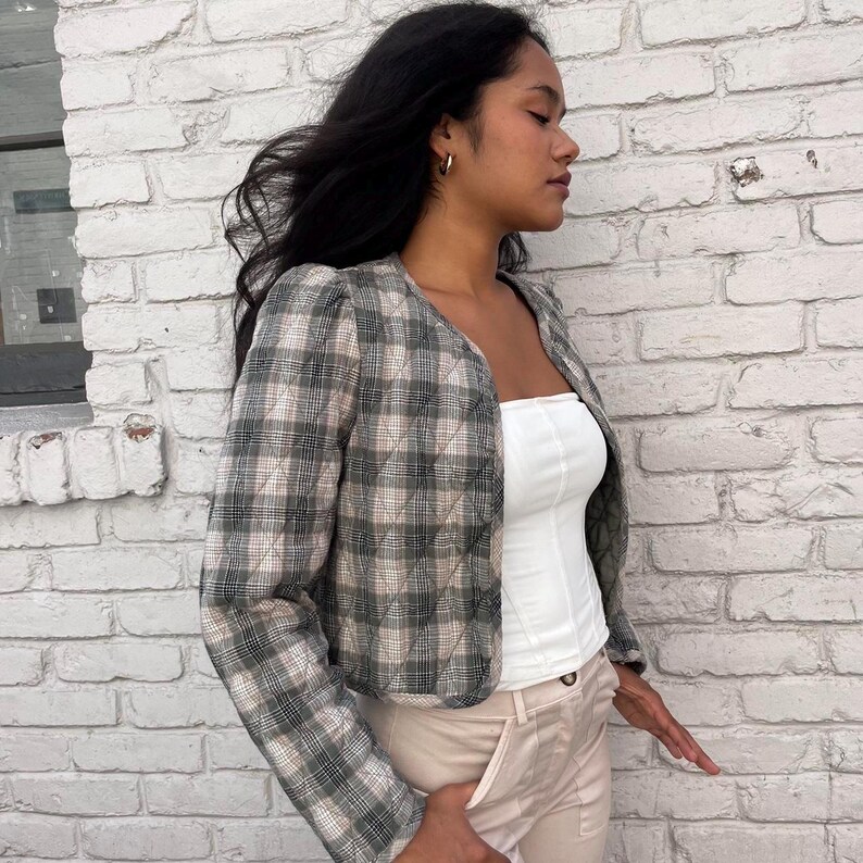 May include: A cropped plaid jacket with a quilted design. The jacket is in a black and white plaid pattern and has a slightly puffed shoulder. The jacket is worn over a white vest top and beige trousers.