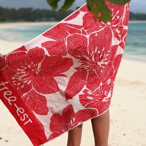 May include: A beach towel with a vibrant red floral pattern on a white background. The towel has a red border with the text "free-est". The towel is being held up on a beach.