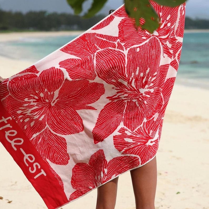 May include: A beach towel with a vibrant red floral pattern on a white background. The towel has a red border with the text "free-est". The towel is being held up on a beach.