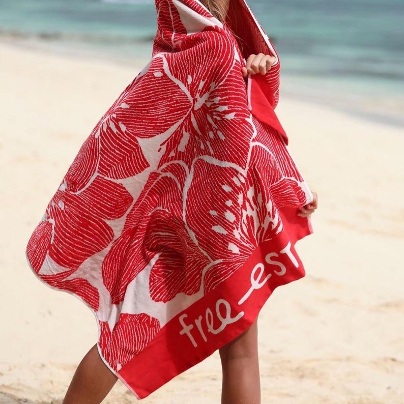 May include: A red and white hooded beach towel with a large floral pattern. The towel has a red border with the text "free-est" in white. The towel is being held up on a sandy beach near the ocean.