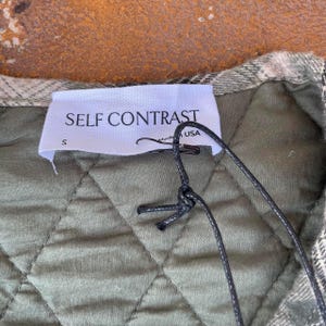May include: A green quilted jacket with a white tag that says "SELF CONTRAST" and "S" in the bottom left corner. The tag is attached to the jacket with a black string.