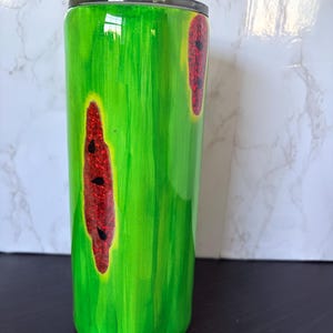 May include: A green tumbler with a watermelon design. The tumbler has a black lid and features a bright green background with two red, glittery watermelon slices with black seeds. The design is painted on the tumbler.
