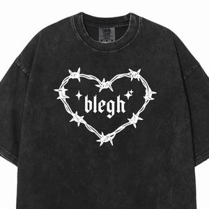 May include: A black acid-wash t-shirt featuring a white graphic print on the chest. The design consists of a heart shape formed by barbed wire with the word blegh written in a gothic font inside, accompanied by two small four-pointed star icons.