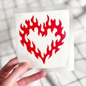 May include: A red flame heart design on a white sticker. The heart shape is formed by stylized flames. The sticker is held against a checkered background.