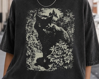 May include: A charcoal gray short-sleeve t-shirt featuring a graphic print of a black cat sitting in a garden with a castle silhouette in the background. The design is rendered in a light cream color, contrasting against the dark fabric of the shirt.