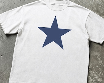 May include: A short-sleeved t-shirt in a light off-white color featuring a large, solid navy blue five-pointed star graphic centered on the chest. The garment has a classic crew neckline and a straight hemline, constructed with a standard fit.
