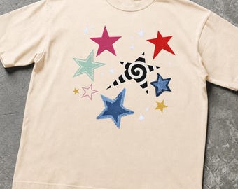 May include: A beige short-sleeve t-shirt featuring a central graphic of multiple stars in various colors and sizes, including a prominent black and white spiral-patterned star, alongside solid red, pink, blue, green, and yellow stars.