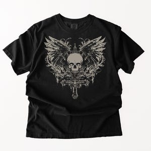 May include: A black short-sleeve t-shirt featuring a large, centered graphic print in a light beige color. The design displays a detailed human skull positioned above an ornate cross, flanked by expansive, feathered wings and decorative flourishes.