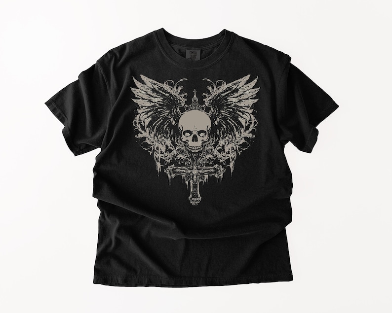 May include: A black short-sleeve t-shirt featuring a large, centered graphic print in a light beige color. The design displays a detailed human skull positioned above an ornate cross, flanked by expansive, feathered wings and decorative flourishes.