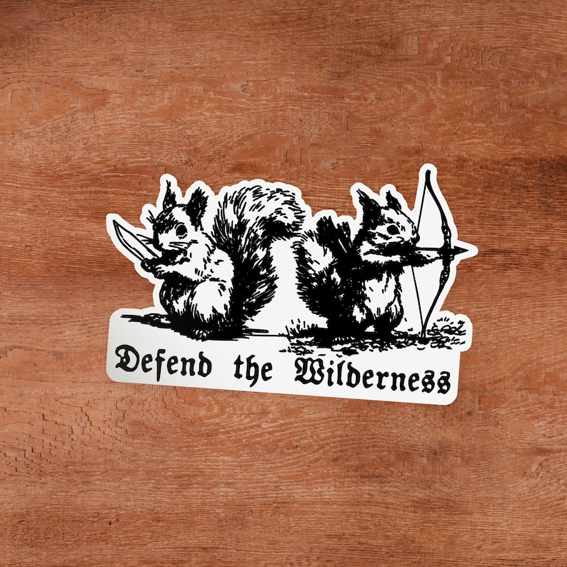 May include: A white sticker featuring a black ink illustration of two squirrels. One squirrel holds a knife, while the other holds a bow and arrow. Below the squirrels, the text reads "Defend the Wilderness" in a black, stylized gothic font.