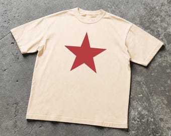 May include: A short-sleeved crew neck t-shirt in a light beige color. The center of the chest features a large, solid red five-pointed star graphic. The garment is constructed with a standard fit, featuring a ribbed neckline and hemmed sleeves.