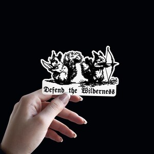 May include: A white sticker with a black ink illustration of two squirrels, one holding a knife and the other holding a bow and arrow. Below the squirrels, the text reads Defend the Wilderness in a stylized black gothic font on a white background.