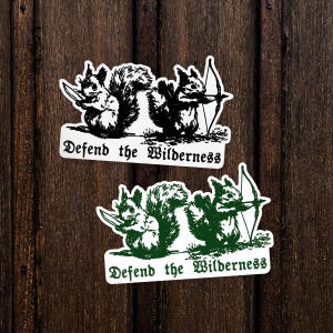 May include: Two rectangular stickers featuring a black and white version and a green and white version. Each sticker depicts two squirrels, one holding a knife and one holding a bow and arrow, with the text Defend the Wilderness below them.