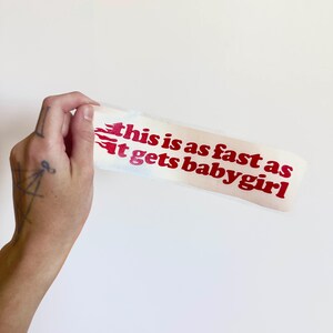 K&ouml;nnte beinhalten: Ein wei&szlig;er Aufkleber mit rotem Text, der "this is as fast as it gets babygirl" lautet. Der Text ist in einer fetten, serifenlosen Schriftart mit Flammengrafiken auf der linken Seite. Der Aufkleber wird vor einem wei&szlig;en Hintergrund gehalten.