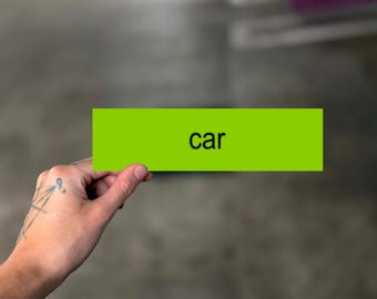 May include: A rectangular lime green card featuring the word car printed in black lowercase letters. The card is held horizontally, displaying a simple design with a solid vibrant green background and clear, bold typography for the text.