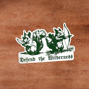 May include: A white sticker with a dark green illustration of two squirrels. One squirrel holds a knife, and the other holds a bow and arrow. Below the squirrels, the text reads "Defend the Wilderness" in a dark green, stylized gothic font.