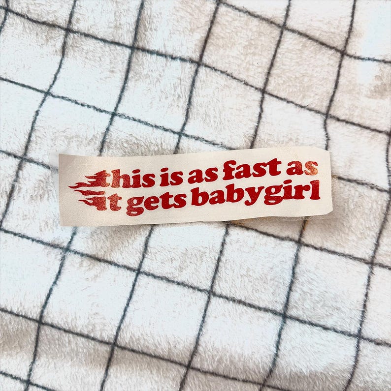K&ouml;nnte beinhalten: Ein rechteckiger Aufkleber mit dem Text "this is as fast as it gets baby girl" in Rot, mit flammenartigen Designs auf der linken Seite. Der Aufkleber befindet sich auf einer wei&szlig;en Oberfl&auml;che mit einem schwarzen Gittermuster.