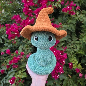 Pu&ograve; includere: Un serpente verde di peluche fatto a mano con un cappello da mago all'uncinetto arancione. Il serpente ha grandi occhi neri e viene tenuto davanti a uno sfondo di fiori rosa e fogliame verde. Il serpente &egrave; fatto di filato morbido e strutturato.
