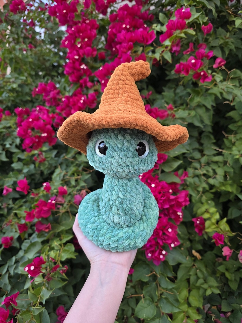 Pu&ograve; includere: Un serpente verde di peluche fatto a mano con un cappello da mago all'uncinetto arancione. Il serpente ha grandi occhi neri e viene tenuto davanti a uno sfondo di fiori rosa e fogliame verde. Il serpente &egrave; fatto di filato morbido e strutturato.
