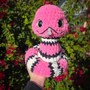 Pu&ograve; includere: Un serpente di peluche rosa fatto a mano con grandi occhi scintillanti e un naso argentato. Il corpo del serpente presenta motivi a strisce bianche e nere. Il testo "Turtles Tangled" &egrave; visibile.