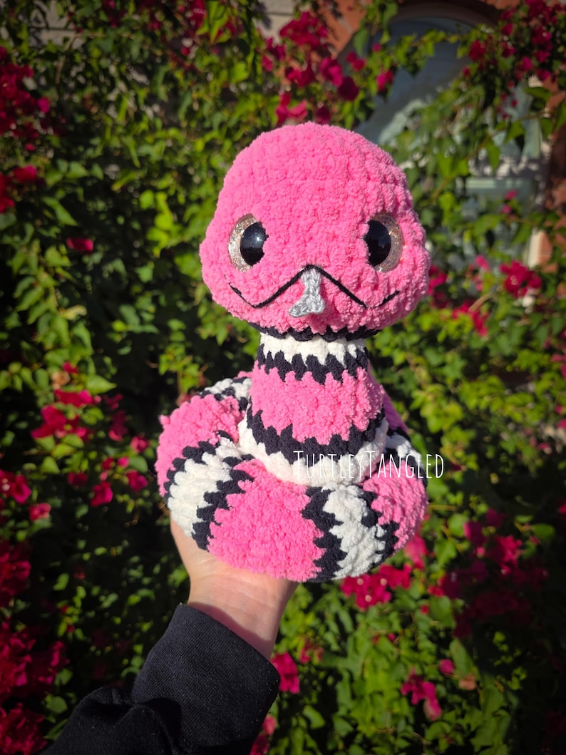 Pu&ograve; includere: Un serpente di peluche rosa fatto a mano con grandi occhi scintillanti e un naso argentato. Il corpo del serpente presenta motivi a strisce bianche e nere. Il testo "Turtles Tangled" &egrave; visibile.