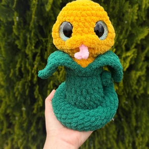 May include: A handmade plush snake toy with a yellow head and a green body. The snake has large, glittery eyes and a pink tongue. The toy is made of soft, textured yarn and is held in a hand.