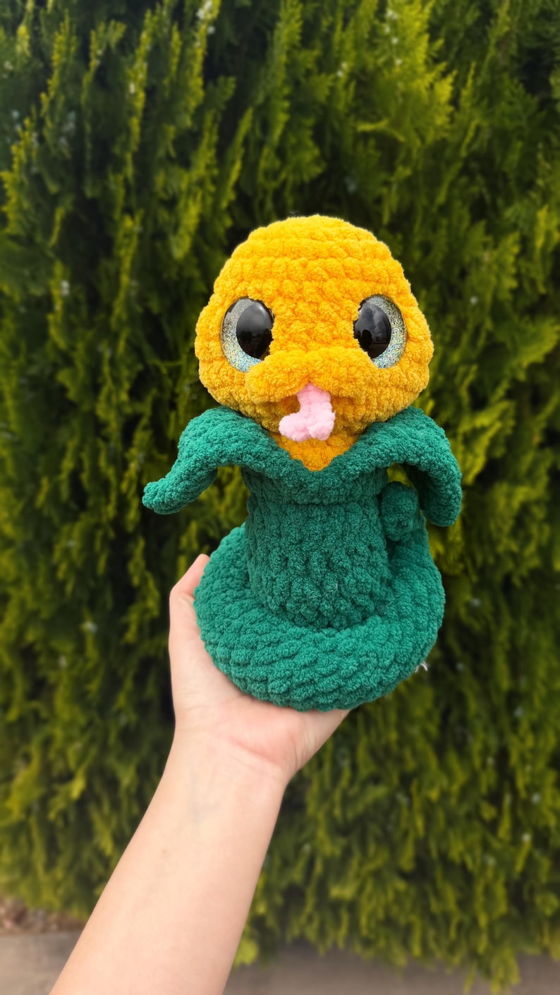 May include: A handmade plush snake toy with a yellow head and a green body. The snake has large, glittery eyes and a pink tongue. The toy is made of soft, textured yarn and is held in a hand.