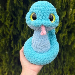May include: A handmade, plush snake toy in shades of blue, with a light pink tongue and large black eyes with yellow accents. The snake is made of a soft, textured material, and is a cute, cuddly toy.