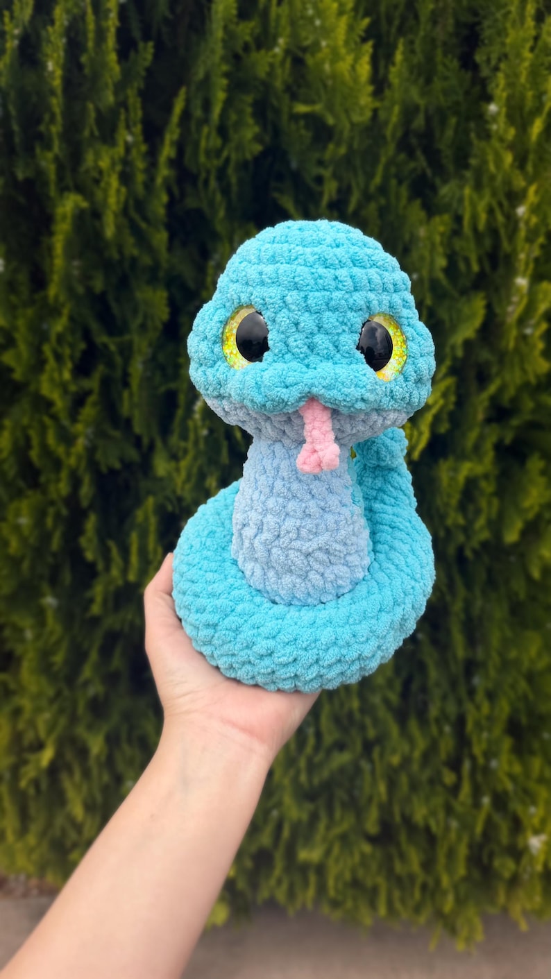 May include: A handmade, plush snake toy in shades of blue, with a light pink tongue and large black eyes with yellow accents. The snake is made of a soft, textured material, and is a cute, cuddly toy.