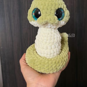 May include: A handmade, plush snake toy in shades of green and cream. The snake has large, glittery blue eyes and a textured, crocheted appearance. The text "TurtleyTangled" is visible on the toy. A QR code is in the lower right corner.
