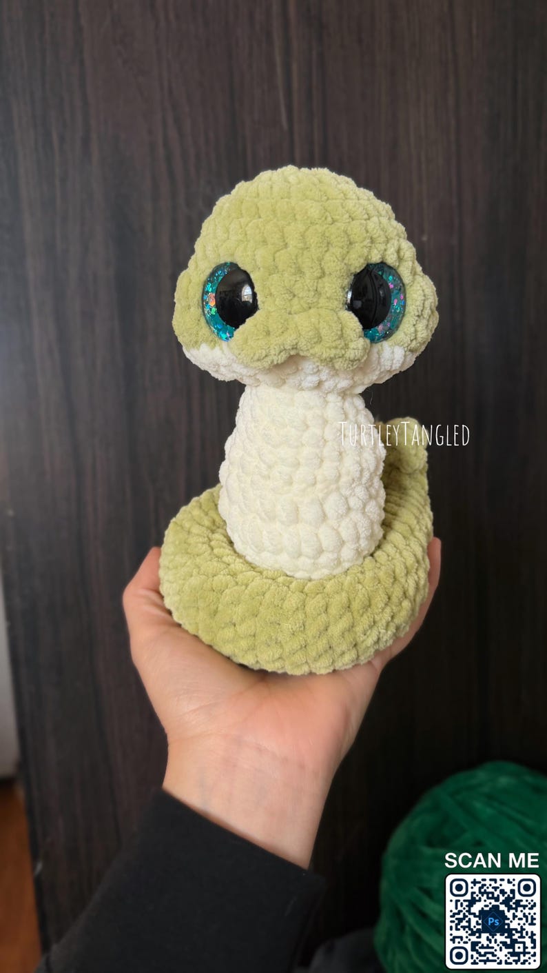 May include: A handmade, plush snake toy in shades of green and cream. The snake has large, glittery blue eyes and a textured, crocheted appearance. The text "TurtleyTangled" is visible on the toy. A QR code is in the lower right corner.