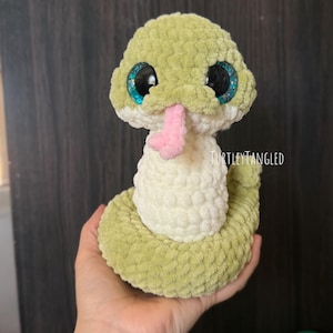 May include: A handmade, plush snake toy in shades of green and white, with large, glittery blue eyes and a pink tongue. The snake is crocheted with a textured yarn, and the text "TurtleyTangled" is visible.