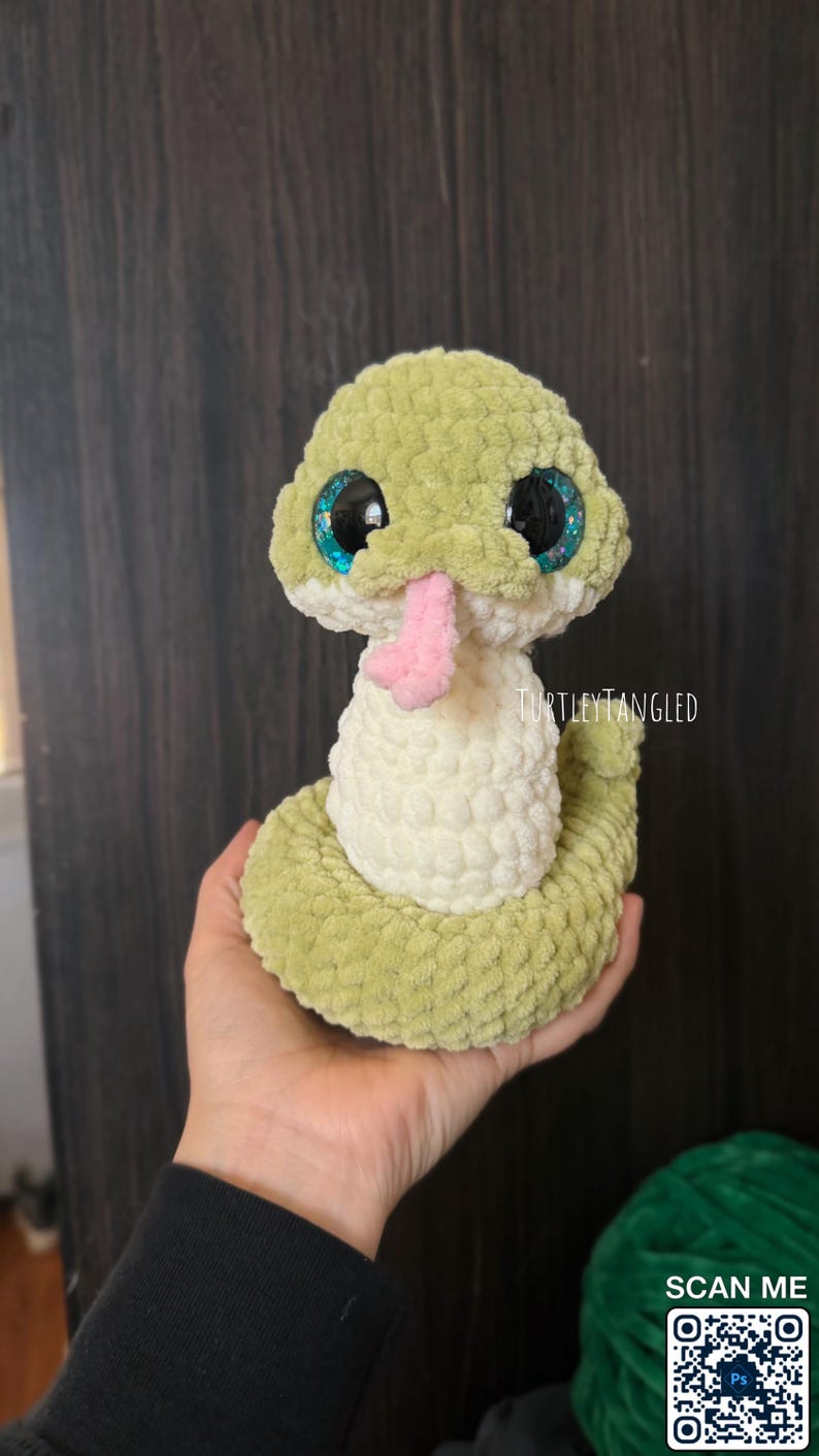 May include: A handmade, plush snake toy in shades of green and white, with large, glittery blue eyes and a pink tongue. The snake is crocheted with a textured yarn, and the text "TurtleyTangled" is visible.