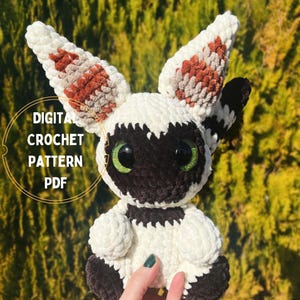 May include: A crocheted plush toy resembling a white and brown creature with large green eyes. The toy is sitting on a hand and has a fluffy texture. The text "DIGITAL CROCHET PATTERN PDF" is visible in the image.