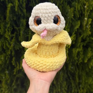 May include: A handmade crochet snake toy in cream and yellow. The snake has large, glittery eyes, a pink tongue, and a yellow body. The toy is soft and plush, perfect for a child's play or nursery decor.
