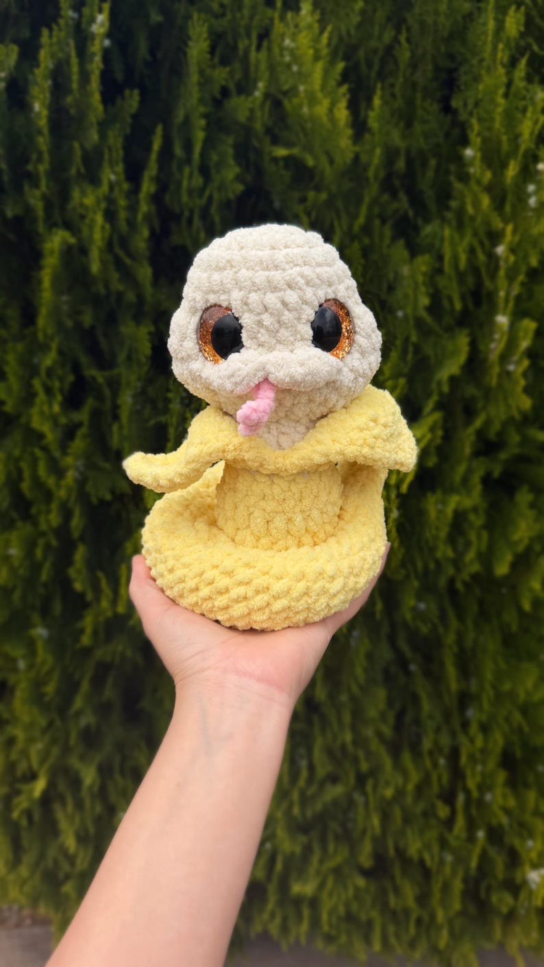 May include: A handmade crochet snake toy in cream and yellow. The snake has large, glittery eyes, a pink tongue, and a yellow body. The toy is soft and plush, perfect for a child's play or nursery decor.
