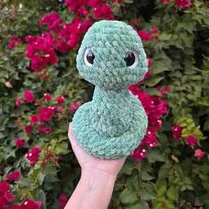 Pu&ograve; includere: Un giocattolo a forma di serpente di peluche verde chiaro fatto a mano con grandi occhi con pupille nere. Il morbido serpente strutturato &egrave; arrotolato e tenuto contro uno sfondo di vibranti fiori rosa e fogliame verde.